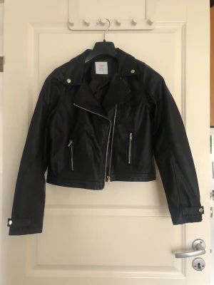 Svart skinnjacka - Snygg svart skinnjacka med klassisk bikerkrage och silvriga dragkedjor framtill samt på fickorna. Är i storlek 164 men passar mig som brukar ha storlek XS/S