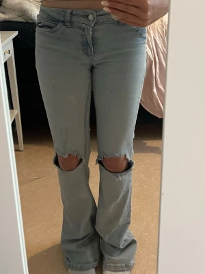 Ljusblå bootcut jeans med hål - Säljer ett par ljusblå bootcut jeans med stora slitningar vid knäna. Jeansen har klassisk femficksmodell, låg midja och vida ben. Perfekta för dig som gillar en avslappnad och trendig stil. Materialet är denim i bomull.