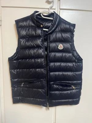 Svart dunväst från Moncler - Snygg svart dunväst från Moncler med klassisk quiltad design och hög krage. Västen har två dragkedjeförsedda fickor framtill och ikonisk Moncler-logga på bröstet. Perfekt för lager-på-lager och streetstyle. Materialet är glansigt och känns lyxigt.