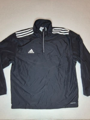 Svart träningsjacka från Adidas - Svart Adidas ClimaProof vindjacka/prasseljacka med klassiska tre ränder på axlarna och halv dragkedja framtill. Tunn, lätt och rymlig modell som passar bra till träning, vardag, sport eller retro/streetwear-look. Storlek UK 46/48, upplevs rymlig i passformen. Märke: Adidas Modell: ClimaProof Storlek: UK 46/48 Färg: Svart/vit Typ: Vindjacka / prasseljacka / träningsjacka Skick: Begagnat skick enligt bilder Mått: 80 cm lång, 67 cm bred, 58 cm ärm mätt från armhåla.