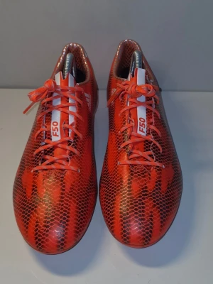 adidas - F50 Adizero FG Röd/Vit/Svart - Säljer ett par Adidas F50 fotbollsskor i rött med svart ormmönster. Skorna har snörning, låg profil och är designade för fart på planen. Ovandelen är i syntetmaterial och sulan har fasta dobbar för spel på gräs. Perfekta för dig som vill sticka ut på planen.