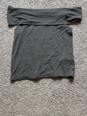 Mörkgrå offshoulder topp Kappahl - Mörkgrå offshoulder topp från Kappahl i mjuk bomullsmix. Enkel och stilren design med bred resår upptill som sitter snyggt över axlarna. Perfekt att matcha med jeans eller kjol för en trendig look.