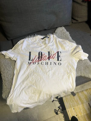 Vit Moschino t-shirt! - Säljer en as fet vit t-shirt från Moschino med tryck i svart och rött framtill. Klassisk passform med korta ärmar och rund halsringning. Perfekt för dig som vill ha en stilren men ändå statement t-shirt, då det står ”Love Moschino Milano”. Väldigt bra skick och letar efter en ny användare, bara att höra av dig vid frågor eller funderingar!