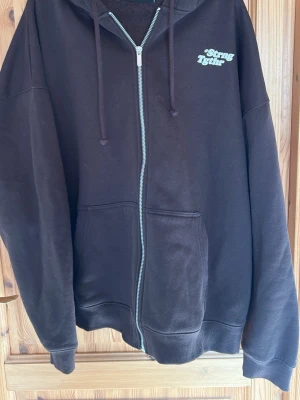 Mörkbrun hoodie med dragkedja och tryck - Mörkbrun hoodie med dragkedja framtill och vit text 'Strong Tydir' på bröstet. Utrustad med huva och dragsnören samt två stora fickor framtill. Tillverkad i mjukt sweatshirtmaterial, perfekt för en avslappnad stil.