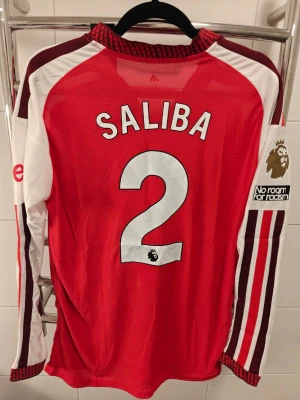 Arsenal nya hemma tröja i spelar version !  långärmad matchtröja Rice 41 - Skaffa den nya hemma tröjan i spelar version. Långärmad Arsenal matchtröja i rött och vitt med Saliba. Tröjan har klubbmärke och sponsortryck framtill, samt klassiska Adidas-detaljer på ärmarna. Tillverkad i lätt och ventilerande funktionsmaterial, perfekt för fotbollsträning eller match.