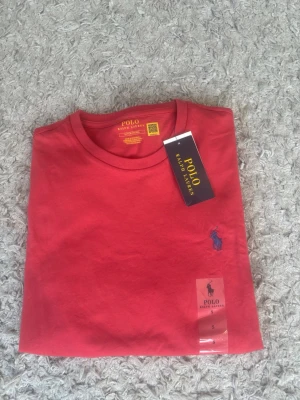 Röd t-shirt från Polo Ralph Lauren - Snygg röd t-shirt från Polo Ralph Lauren med klassisk blå logga broderad på bröstet. Tillverkad i mjuk bomull och har en normal passform med korta ärmar och rund hals. Perfekt till jeans eller shorts för en clean look.