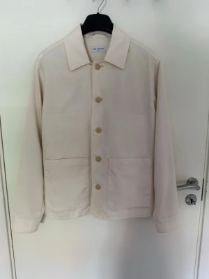 Klassisk beige overshirt i bomull - Stilren beige overshirt med stora framfickor, Skjortkrage och knäppning med fyra knappar framtill från these glory days. Tillverkad i ett mjukt bomullstyg som ger en avslappnad men ändå snygg look. Perfekt att bära över en t-shirt eller skjorta för mer formella event.