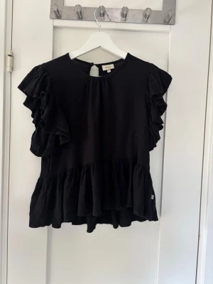 Svart volangblus med kort ärm - Säljer en svart blus med volangdetaljer och korta, puffiga ärmar. Blusen har rund halsringning och en lös passform med volang nedtill.💗 storlek 145/152