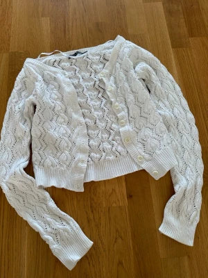 Vit stickad kofta från Soul River - Snygg vit stickad kofta från Soul River i storlek S. Koftan har ett vågigt hålmönster, långa ärmar och ribbade muddar. Perfekt att slänga över en topp för en chill look. Stängs med vita knappar framtill.