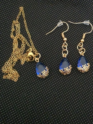Guldhalsband med mörkblå droppe - Snyggt halsband med tunn guldfärgad kedja och en droppformad mörkblå sten infattad i guldfärgad metall med blommönster. Matchar perfekt med örhängena i samma stil för en elegant look. Materialet är metall och glasliknande sten.