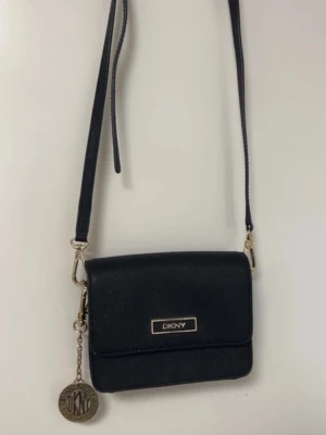 Mini Svart axelväska från DKNY - Snygg svart axelväska från DKNY i stilren design med guldfärgade detaljer och logotyp framtill. Väskan har en justerbar axelrem och ett dekorativt DKNY-mynt som hänger på sidan. Perfekt storlek för det viktigaste och tillverkad i ett slätt, tåligt material.