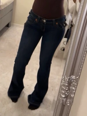 True religion Joey jeans  - Snygga mörkblå bootcut jeans med kontrastsömmar och dekorativa fickor bak. Jeansen har låg midja och klassisk femficksdesign. Materialet är stretchigt denim som sitter bekvämt och formar sig efter kroppen. Perfekta för en trendig och avslappnad look.