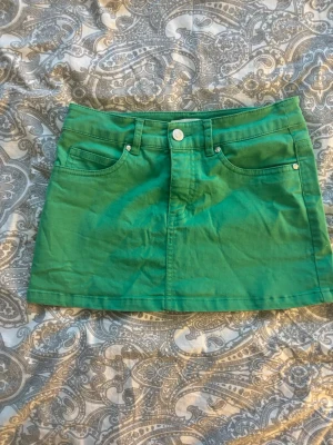 Lågmidjad grön kortkjol från att. - Snygg lågmidjad grön kortkjol från att. i klassisk jeansmodell med fem fickor och knapp- samt dragkedjestängning. Kjolen är tillverkad i ett mjukt bomullsmaterial och har bälteshällor i midjan. Perfekt för dig som vill ha en färgklick i garderoben!