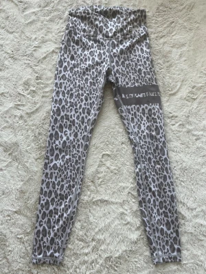 Leopard träningsbyxor - Leopardmönstrade leggings i grå och vita toner med hög midja och bred resår. Materialet är stretchigt och följsamt, perfekt för dig som gillar att sticka ut. På ena benet finns en dekorativ rand med textdetalj som ger extra attityd.
