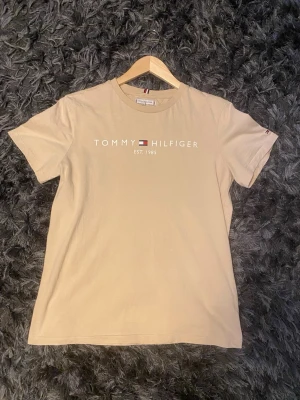 Beige T-shirt från Tommy Hilfiger - Snygg beige T-shirt från Tommy Hilfiger med klassisk logga och texten 'EST. 1985' på bröstet. T-shirten har rund halsringning och korta ärmar. Perfekt basic-plagg med stilren design och mjukt bomullsmaterial.