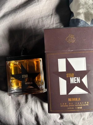 Star Men Nebula Eau de Parfum - Star Men Nebula Eau de Parfum med en stilren, fyrkantig glasflaska och svart stjärndesign. Doften kommer i en elegant brun och guld kartong med tydlig stjärnlogga. Flaskan har svart spraymunstycke och rymmer 100 ml.