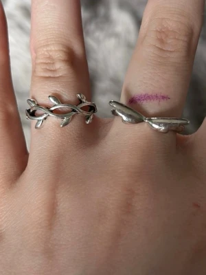 Två silvriga ringar med bladmotiv - Säljer två snygga ringar i silverfärgad metall, båda med bladliknande design. Den ena ringen har ett öppet, flätat mönster av små blad medan den andra har en mer solid form med blad på ovansidan. Perfekta för dig som gillar naturinspirerade accessoarer.
