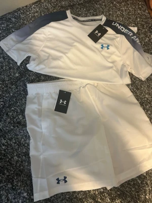 Vitt set från Under Armour - Säljer ett fräscht vitt sportset från Under Armour med t-shirt och shorts.  Settet är i storlek S och sitter riktigt bra. T-shirten har gråa detaljer på axlarna och logga på bröstet, medan shortsen har elastisk midja och logga på benet. Perfekt för träning eller sport. Materialet är lätt och andas. Den är helt ny och oanvänd!!! 