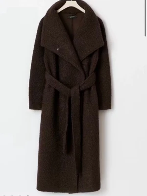 Mörkbrun teddy kappa med skärp - Lång, mörkbrun teddy kappa med bred krage och knytband i midjan. Kappan har en oversized passform och är gjord i ett fluffigt material som ger en trendig look. Perfekt för kalla dagar när du vill vara både varm och snygg.