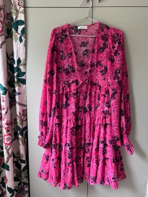 Rosa blommig volangklänning - Supersöt kort klänning i stark rosa med svart blommönster. Klänningen har v-ringning, långa ärmar med resår vid handlederna och flera volanglager. Lite transparent fint blommigt tyg.