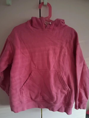 Rosa hoodie med huva och magficka - Säljer en mysig rosa hoodie med stor huva och klassisk magficka framtill. Tröjan har ett litet broderat tryck på bröstet och är gjord i mjukt fleecematerial. Perfekt för chill dagar eller när du vill vara extra bekväm. I använt skick säljes billigt