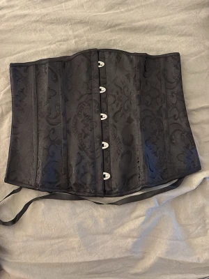 Svart mönstrad satin-korsett - Snygg svart korsett i satin med diskret blommönster. Framsidan har metallknäppning och baksidan snörning med breda satinband för justerbar passform. Korsetten har markerade sömmar och ger en klassisk siluett. Perfekt för dig som vill ha en elegant och edgy look.