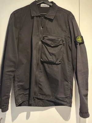 Svart overshirt från Stone Island - Snygg svart overshirt från Stone Island med dragkedja framtill, stor bröstficka med knapp och den klassiska logotypen på vänster ärm. Tillverkad i ett slitstarkt bomullsmaterial och har en avslappnad passform. Perfekt för lager på lager och streetwear-stil.