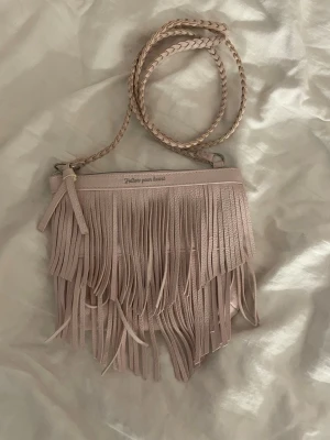 Ljusrosa axelväska med fransar - Supersöt ljusrosa axelväska i fejkläder med långa fransar framtill och flätat axelband. På väskan står det 'Follow your heart' i silvertext. Perfekt för dig som gillar boho-stil och vill sticka ut med en unik accessoar.