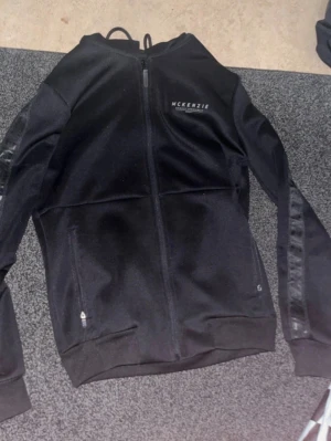 Svart McKenzie hoodie  - Snygg svart hoodie från McKenzie med dragkedja framtill och diskret logga på bröstet. Ärmarna har en blank detalj med text längs sidorna. Tillverkad i ett mjukt och stretchigt material som känns bekvämt. Perfekt för en avslappnad streetwear-look.