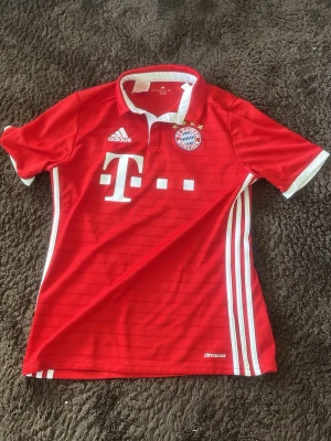 Bayern München matchtröja röd - Säljer en röd Bayern München matchtröja från Adidas med vita detaljer, klubbmärke och sponsortryck på bröstet. Nummer 35 och Sanches på ryggen. Klassisk pikékrage och korta ärmar. Tillverkad i lätt och ventilerande material, perfekt för fotbollsträning eller match.