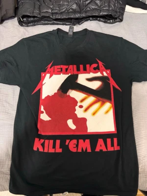 Metallica Kill 'Em All t-shirt svart - Svart t-shirt från Gildan med Metallica Kill 'Em All-tryck i rött, vitt och gult framtill. Klassisk passform med korta ärmar och rund hals. Tillverkad i kraftig bomull, perfekt för dig som gillar bandmerch och rockstil. Passar S