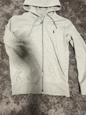 Ljusgrå hoodie från Polo Ralph Lauren - Snygg äkta ljusgrå hoodie från Polo Ralph Lauren med dragkedja framtill och klassisk huva. Broderad logga på bröstet och två fickor på sidorna. Perfekt för en chill och stilren look. Tillverkad i mjukt bomullsmaterial.