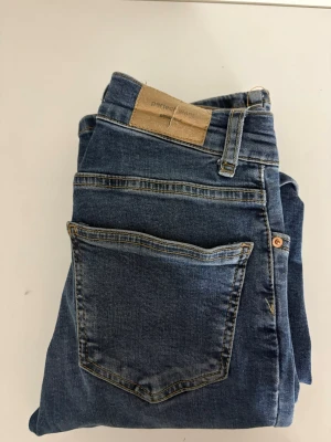 Blå bootcut jeans med fickor - Säljer ett par klassiska blå bootcut jeans med fem fickor och normalhög midja. Jeansen har en snygg tvätt och utsvängda ben, perfekta för en trendig look. Materialet är denim i bomull med lite stretch för skön passform.