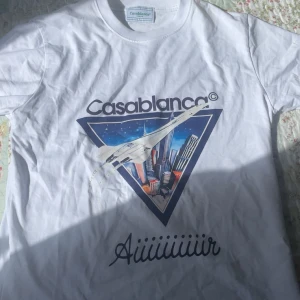 Vit Casablanca t-shirt med Concorde-tryck - Snygg vit t-shirt från Casablanca med stort grafiskt tryck framtill som visar ett Concorde-plan och stadssilhuett. Klassisk rund hals och korta ärmar. Materialet är mjuk bomull och passformen är normal. Perfekt statement-plagg för dig som gillar unika prints.