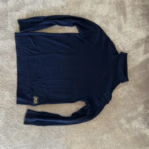 Mörkblå polotröja Scotch & Soda - Snygg mörkblå polotröja från Scotch & Soda med ribbade muddar och hög krage. Tröjan har en klassisk passform och är tillverkad i mjukt material, perfekt för kyligare dagar. Diskret logga nertill på framsidan. 