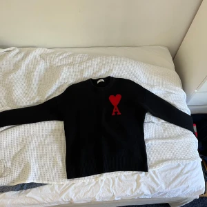 Svart stickad tröja från Ami Paris - Svart stickad tröja från Ami Paris i storlek L. Tröjan har ett rött hjärta med bokstaven A broderat på bröstet, rund hals och ribbade muddar vid ärmslut och nederkant. Perfekt för dig som gillar stilrena plagg med en unik detalj.