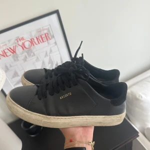 Svarta sneakers från Axel Arigato - Svarta sneakers från Axel Arigato med klassisk låg siluett och vit platt sula. Skorna har svart snörning, rund tå och detaljer i svart mocka vid hälen. Materialet är skinn med en stilren design och guldfärgad logga på sidan.