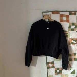 Svart croppad sweatshirt från Nike - Svart croppad sweatshirt från Nike med klassisk vit logga broderad på bröstet. Tröjan har rund hals, långa ärmar och ribbade muddar. Materialet är mjuk bomull med lite stretch för extra komfort.