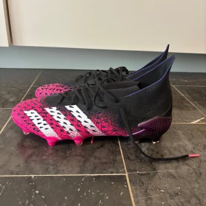 Adidas Predator fotbollsskor - Säljer ett par Adidas fotbollsskor med grymt cool design i rosa och svart med vita ränder på sidorna. Skorna har snörning, mönstrad ovandel och platt sula med dobbar för bästa grepp på planen. Materialet är syntet och skon har en tight passform med hög krage.