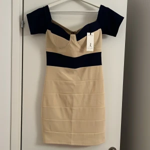Beige och marinblå kort klänning R - Snygg kort klänning från Rare London i beige med marinblå detaljer. Klänningen har offshoulder-ärmar, markerad byst och en bred marinblå rand över bröstet. Baktill finns en dragkedja som gör den enkel att ta på. Perfekt för dig som gillar stilrena kontraster. Med prislapp på helt ny.