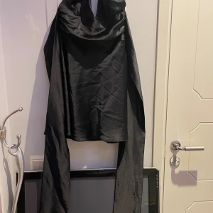 Svart klänning med halterneck som knyts bak i ryggen - Snygg svart klänning med halterneck och långa band som kan knytas i ryggen. Klänningen har en draperad framsida och är tillverkad i ett glansigt, mjukt material som ger en lyxig känsla. Perfekt för dig som vill ha en stilren och elegant look. Lite repig vid kragen. 