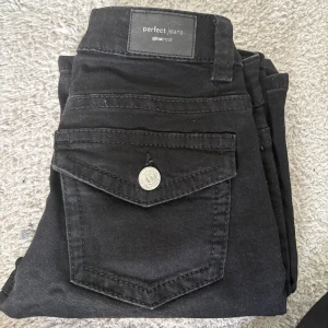Svarta bootcut jeans från Gina Tricot - Säljer ett par svarta bootcut jeans från Gina Tricot, modell Perfect Jeans. Jeansen har låg midja, klassiska fem fickor och snygga silverknappar på bakfickorna. Materialet är stretchigt och sitter riktigt bra på kroppen.
