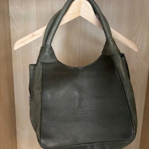 Grön axelväska i skinn från T. Herman - Snygg grön axelväska från T. Herman i mjukt skinn. Väskan har rundad form, breda handtag och en praktisk dragkedja på baksidan. Perfekt storlek för att få plats med allt du behöver och stilren logga framtill. Priset kan diskuteras!