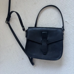 Svart axelväska i skinn - Stilren svart axelväska i skinn med justerbar rem och snyggt spänne framtill. Perfekt storlek för det viktigaste och passar till många olika outfits. Enkel och klassisk design som funkar året runt.