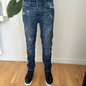 Blå skinny jeans från Jack & Jones - Säljer ett par blå skinny jeans från Jack & Jones med slitna detaljer och snygg tvätt. Jeansen har klassisk femficksdesign, normal midja och smal passform. Tillverkade i bomull med lite stretch för extra komfort.