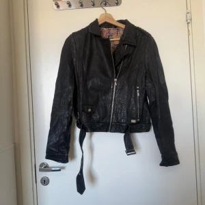 Svart skinnjacka från Jofama, stl 40 - Snygg svart skinnjacka från Jofama med coola detaljer som bälte i midjan, dragkedjor och fickor. Jackan har blommigt foder och klassisk bikerstil med krage och långa ärmar. Perfekt för dig som vill ha en edgy look. Storlek 40, men dessa är känd att vara små i storlek.  Den är i superfin skick, men kan ha vanliga tecken på användning. Finns vanliga tecken som en skinnjacka brukar få. 