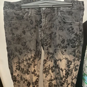 Grå blommiga jeans från H&M, 30/32 - Snygga grå jeans från H&M med svart blommigt mönster över hela byxan. Klassisk femficksmodell i denim med normal passform och raka ben. Perfekta för dig som vill sticka ut med en unik look. Blommorna är mjuka och fluffiga.
