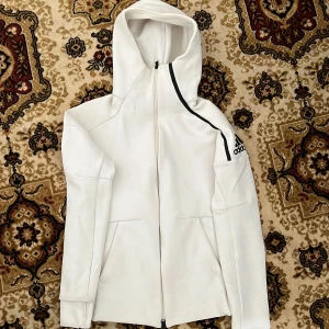 vit hoodie från Adidas med hel dragkedja  - Snygg vit hoodie från Adidas med hel dragkedja och stor huva. Tröjan har svarta detaljer på axeln och Adidas-logga på ärmen. Perfekt för träning eller chill, med två fickor framtill och lång ärm. Materialet är mjukt och känns sportigt.