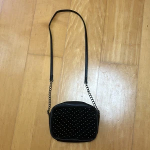 Svart axelväska med nitar och kedja - Säljer en svart axelväska i syntetmaterial med fyrkantig form. Väskan har en lång axelrem i kombination av kedja och läderimitation samt dragkedja upptill. Framsidan är dekorerad med små silvriga nitar i ett prickigt mönster. Perfekt för att lyfta din outfit.
