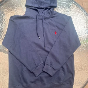 Mörkblå hoodie från Polo Ralph Lauren - Snygg mörkblå hoodie från Polo Ralph Lauren med klassisk röd broderad logga på bröstet. Tröjan har huva med snörning, känguruficka och långa ärmar. Tillverkad i mjuk bomull för en skön och avslappnad stil. Det är XL men mycket liten i storleken är som en M